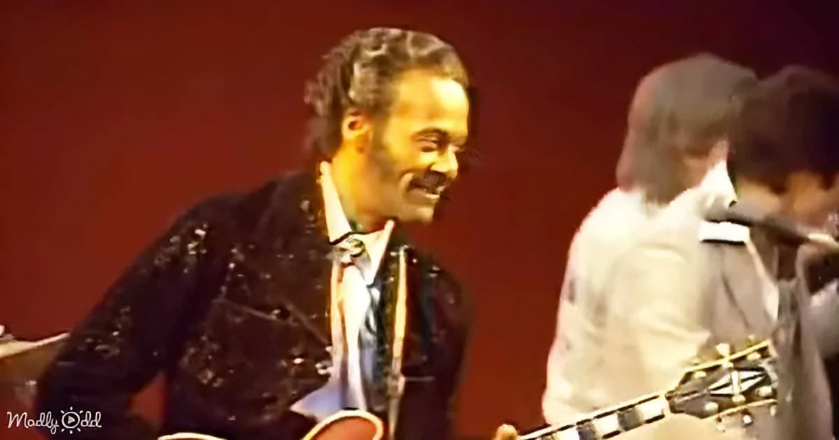 A man with dreadlocks and a mustache plays guitar on stage, smiling, with other musicians in the background.
