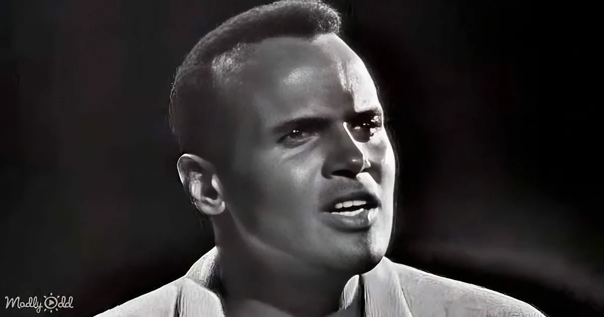 A man in a light-colored shirt appears to be speaking or singing, with a dark background.