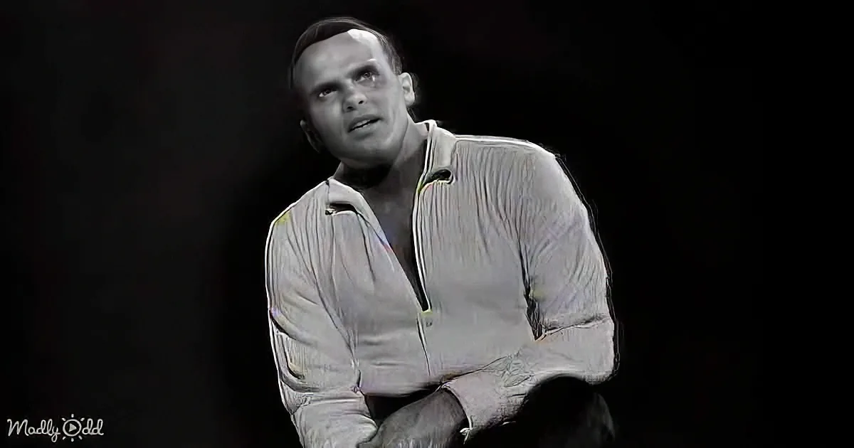 A man in a white, textured jacket sits with his arms crossed, looking upwards against a dark background.