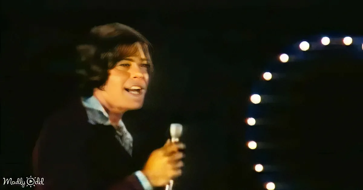 A person with long hair, wearing a suit, sings into a microphone on stage with a circular light backdrop.