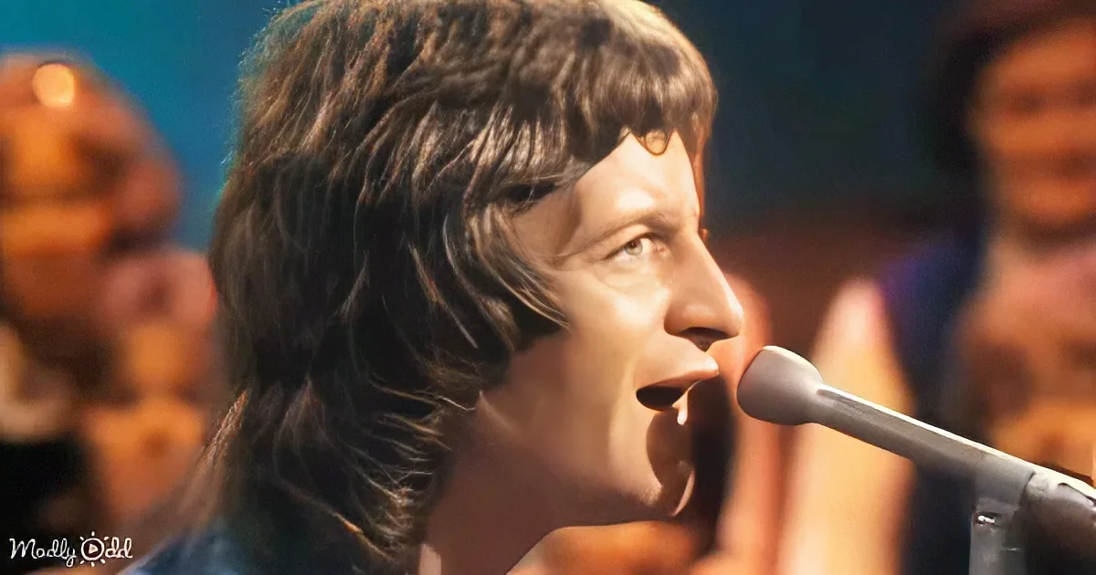 A man with long, dark hair singing into a microphone, with a blurred background.