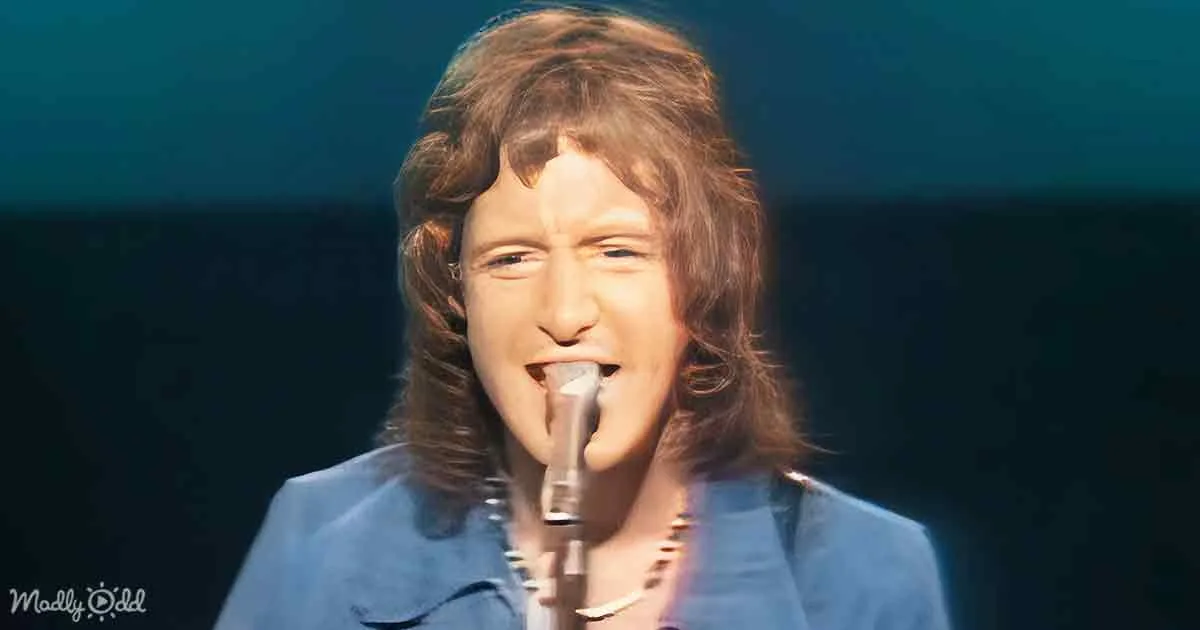 A person with long, wavy hair sings into a microphone, wearing a blue shirt, against a dark background.