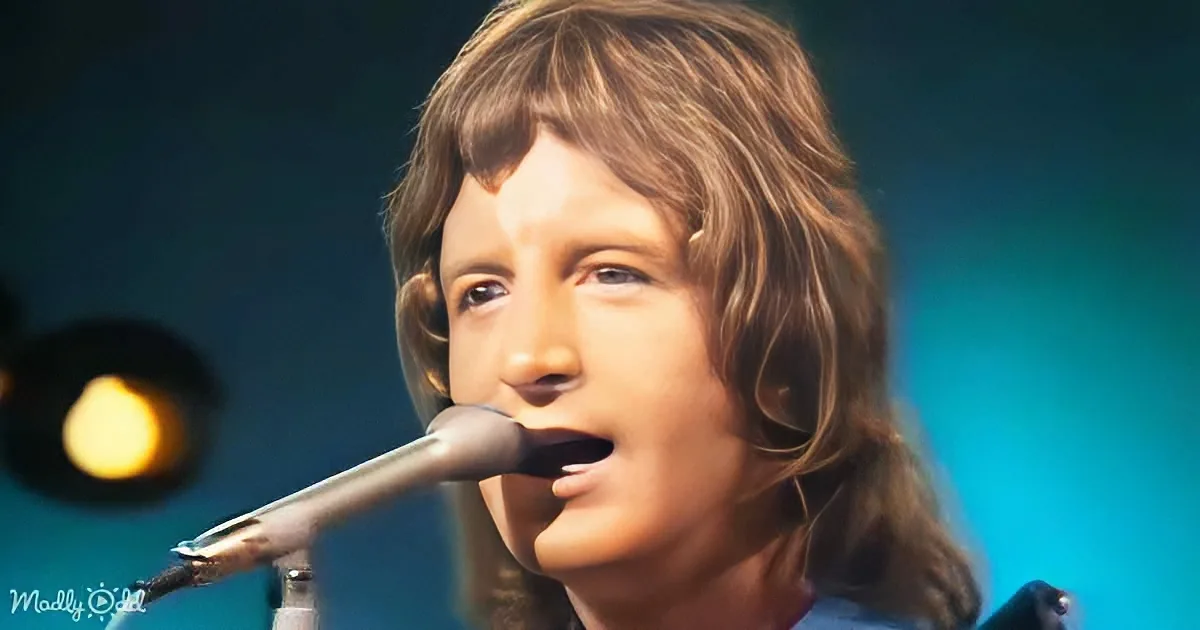 A person with shoulder-length brown hair sings into a microphone against a dark background with a blue light.