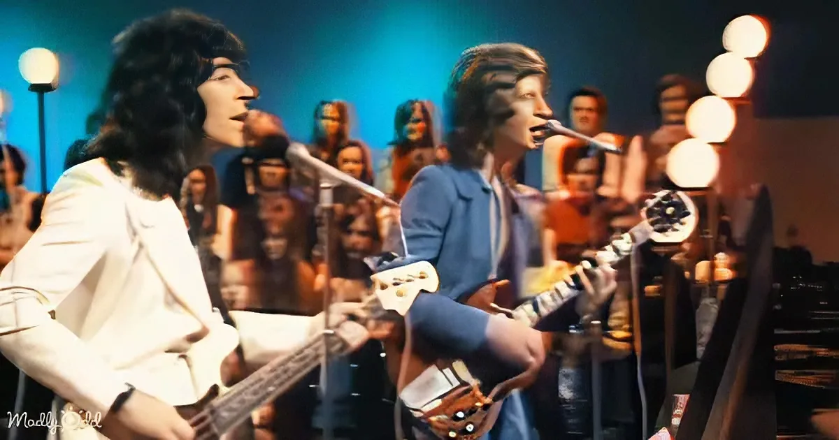 Two musicians, one in white, the other in blue, play guitars on stage with a crowd and bright lights in the background.