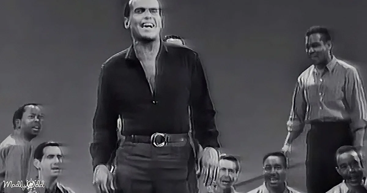 A man in a black shirt and jeans stands confidently, surrounded by a crowd, in a black-and-white image.
