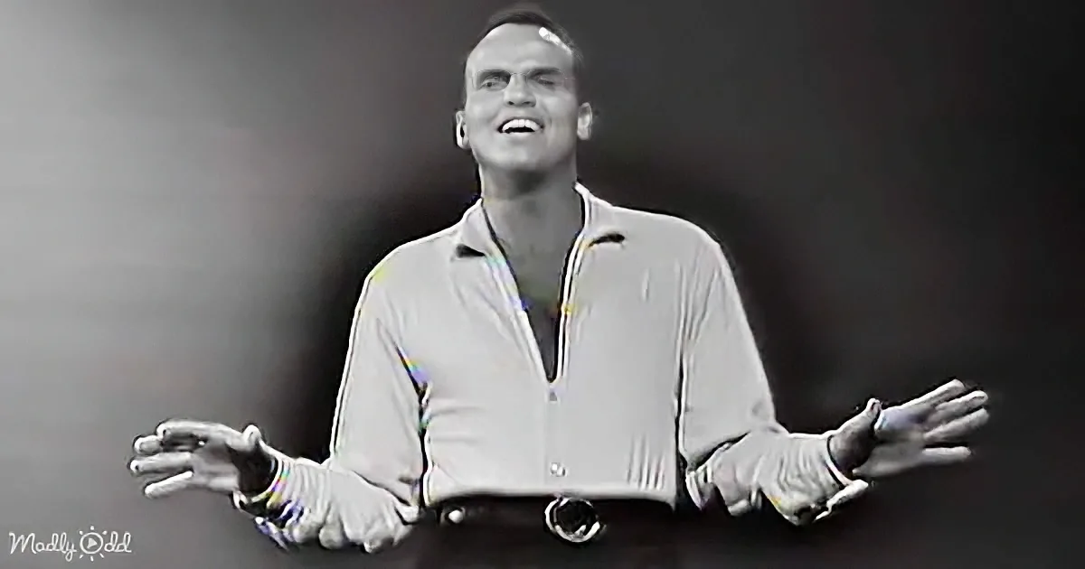 A man in a light-colored shirt and dark pants stands with arms outstretched, smiling, against a dark background.