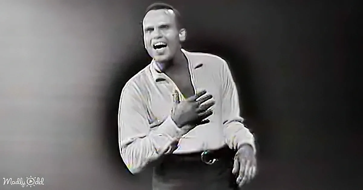 A man in a light-colored shirt sings passionately, hands clasped near his chest.