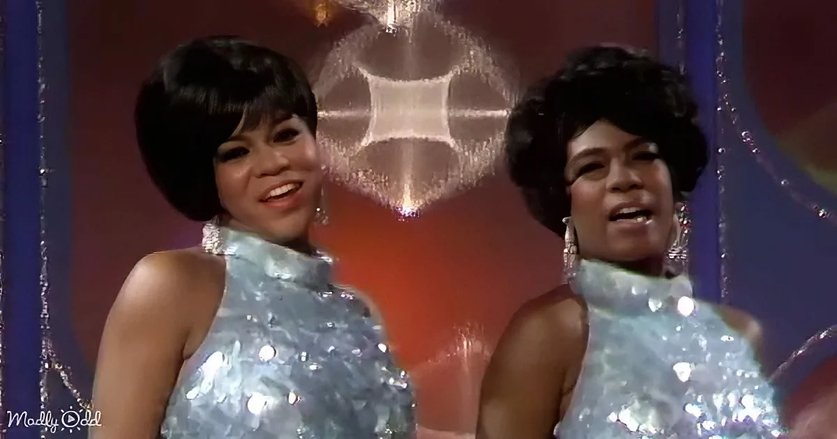 Two women in shimmering silver dresses with large earrings, standing in front of a reflective backdrop.