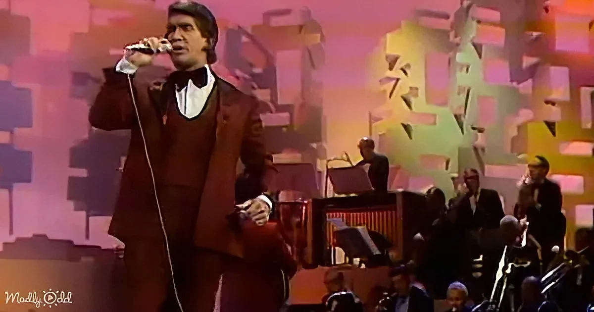 A man in a brown suit sings into a microphone on stage with a band and colorful backdrop.