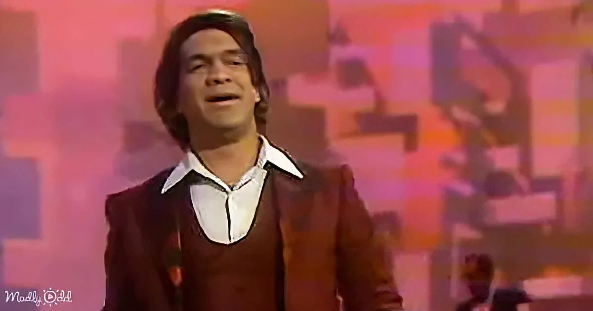 A man in a brown suit and white shirt sings on stage with a colorful background.
