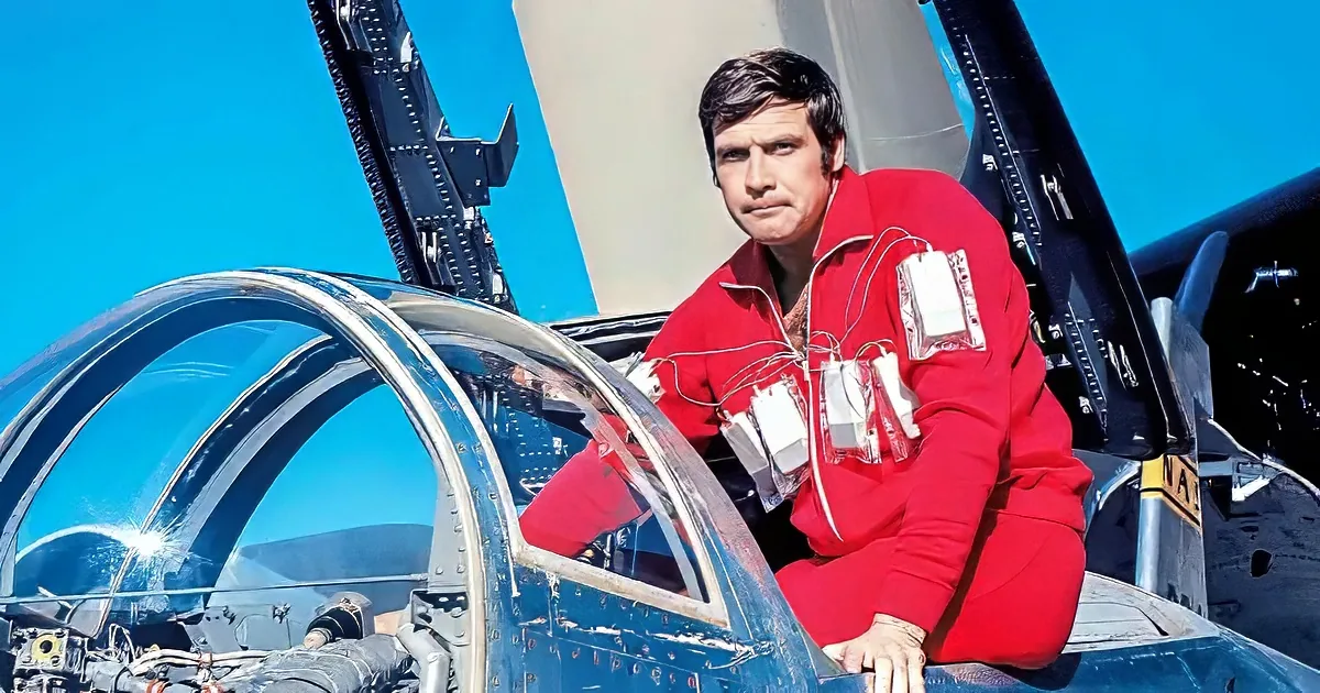 A man in a red jumpsuit with sensors sits in the cockpit of a jet, with a clear blue sky in the background.