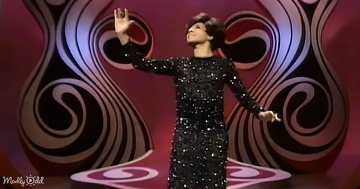 A person in a sparkly black dress stands on stage with a vibrant, swirling background.
