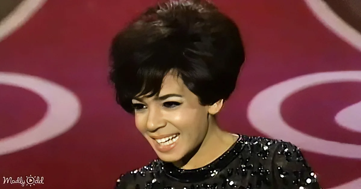 A smiling woman with a short, styled hairstyle and a sequined black top against a red background.