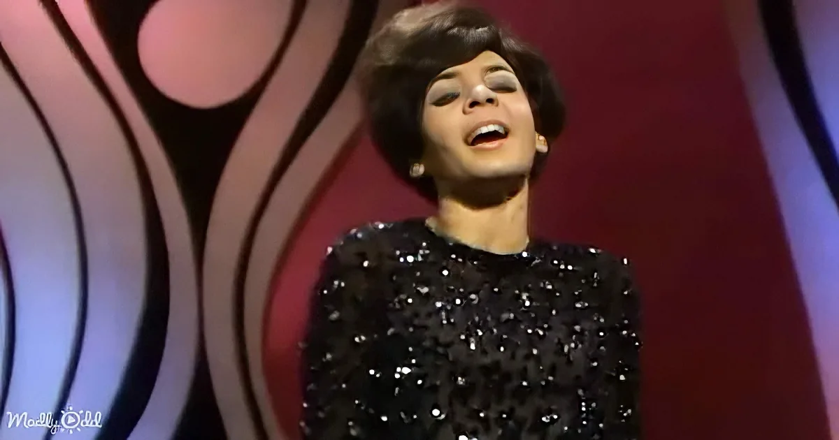 A woman in a black sequined dress sings on stage with a vibrant, abstract background.