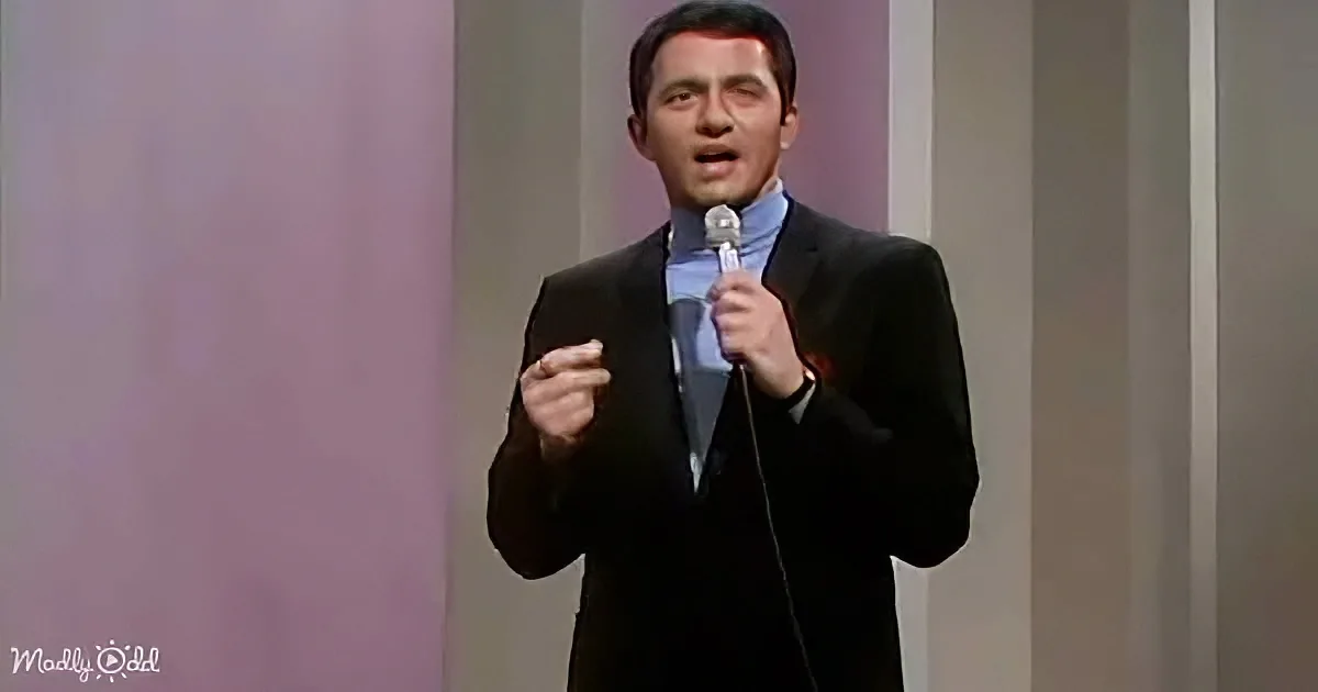 A man in a black suit holds a microphone, speaking on stage with a purple background.