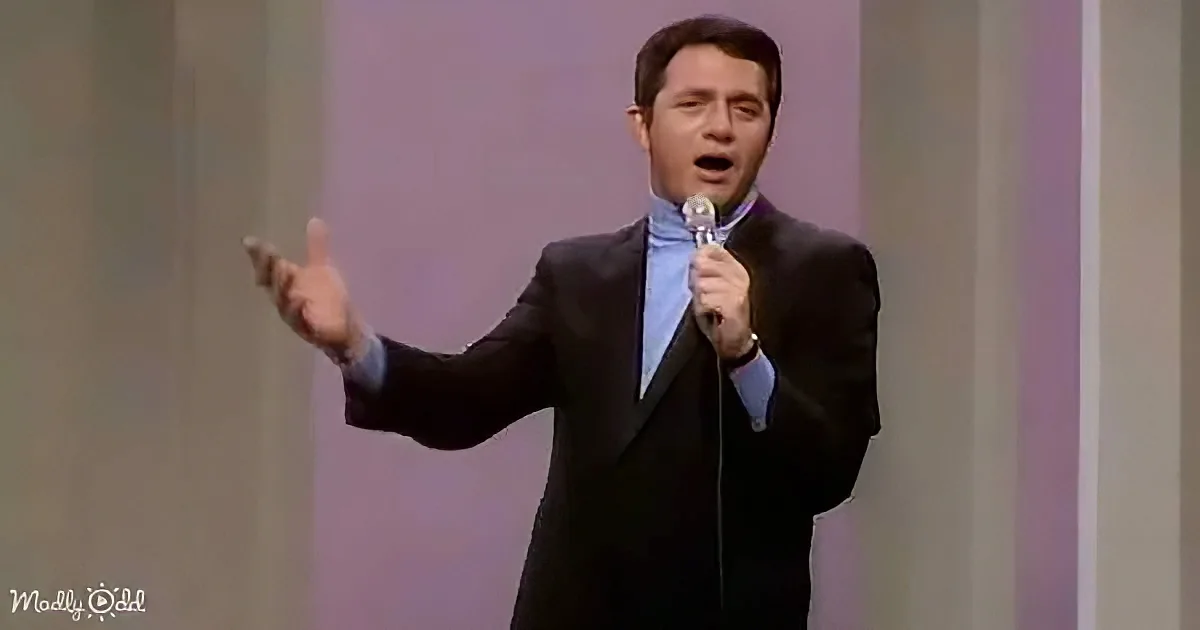 A man in a suit holds a microphone, gesturing with his right hand, possibly singing or speaking on stage.
