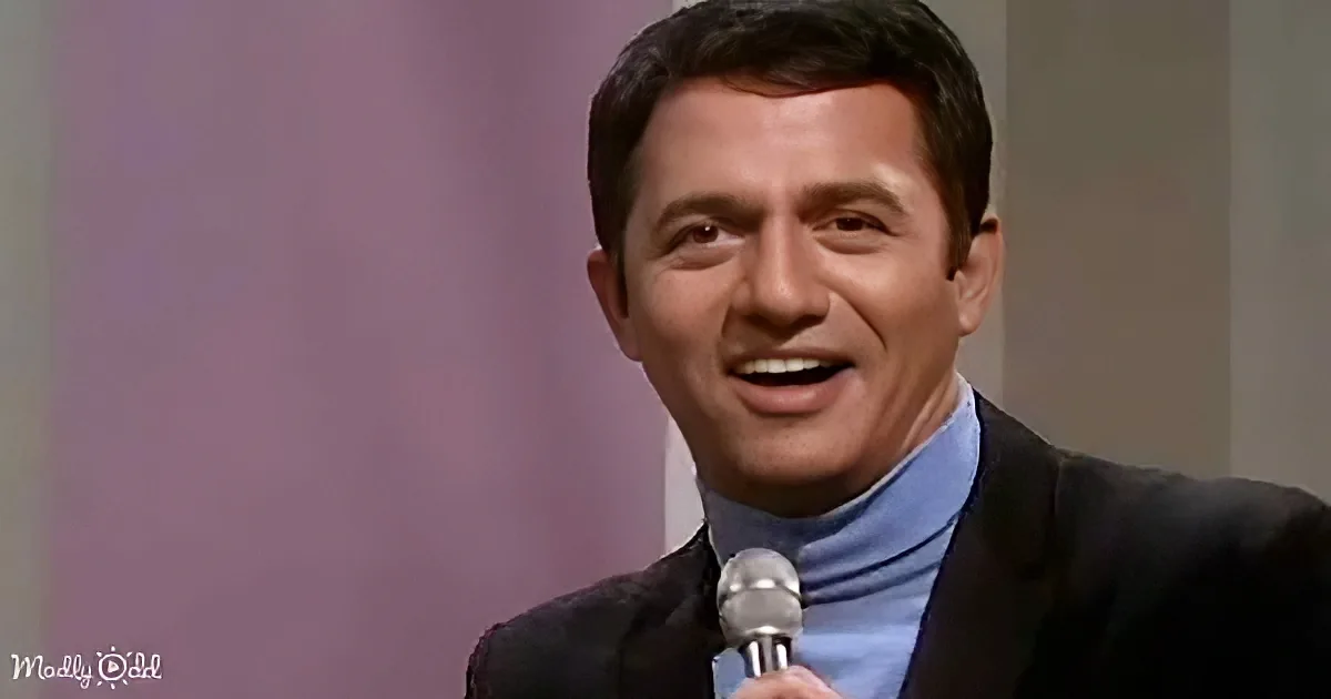A man in a suit and turtleneck smiles while holding a microphone.
