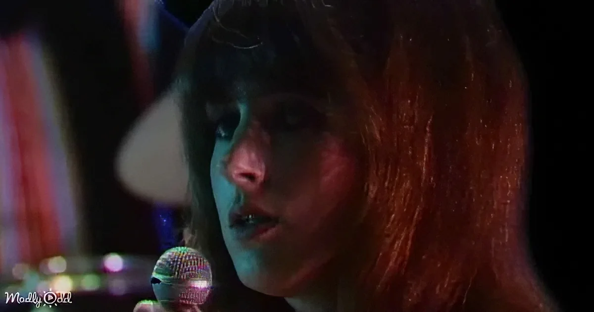 A person with long, reddish-brown hair holds a microphone, illuminated by stage lighting.