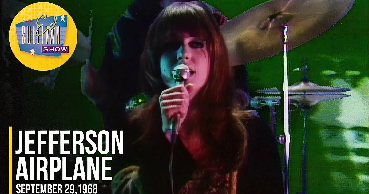 A person with red hair and bangs sings into a microphone, surrounded by a dark, blurred background.