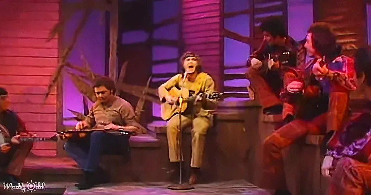 A man plays guitar and sings on stage, surrounded by musicians and a colorful backdrop.