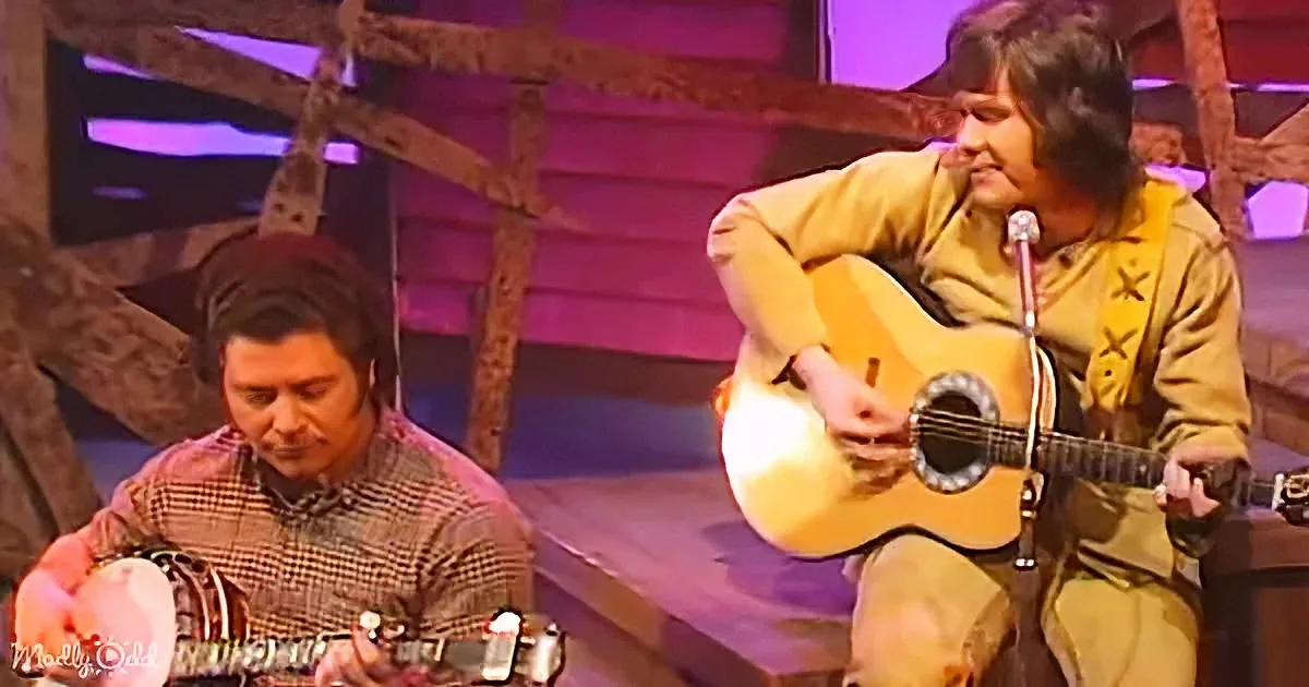 A man in a yellow outfit plays guitar on stage, another man in a plaid shirt plays banjo.
