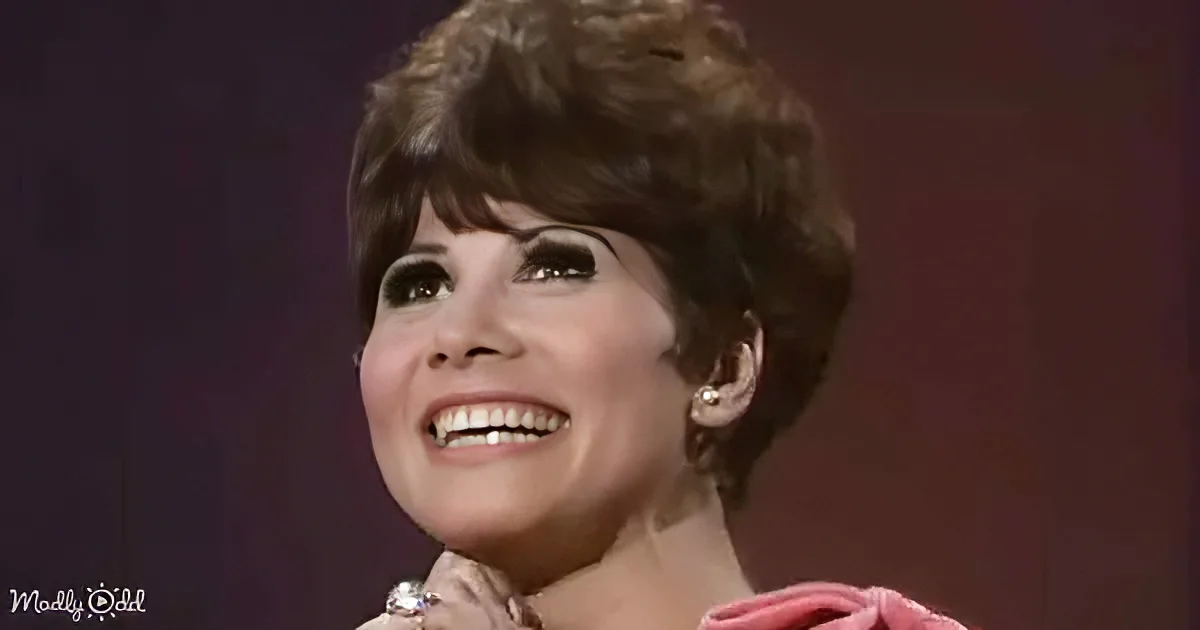 A smiling woman with short brown hair, wearing a pink top and earrings, against a dark background.