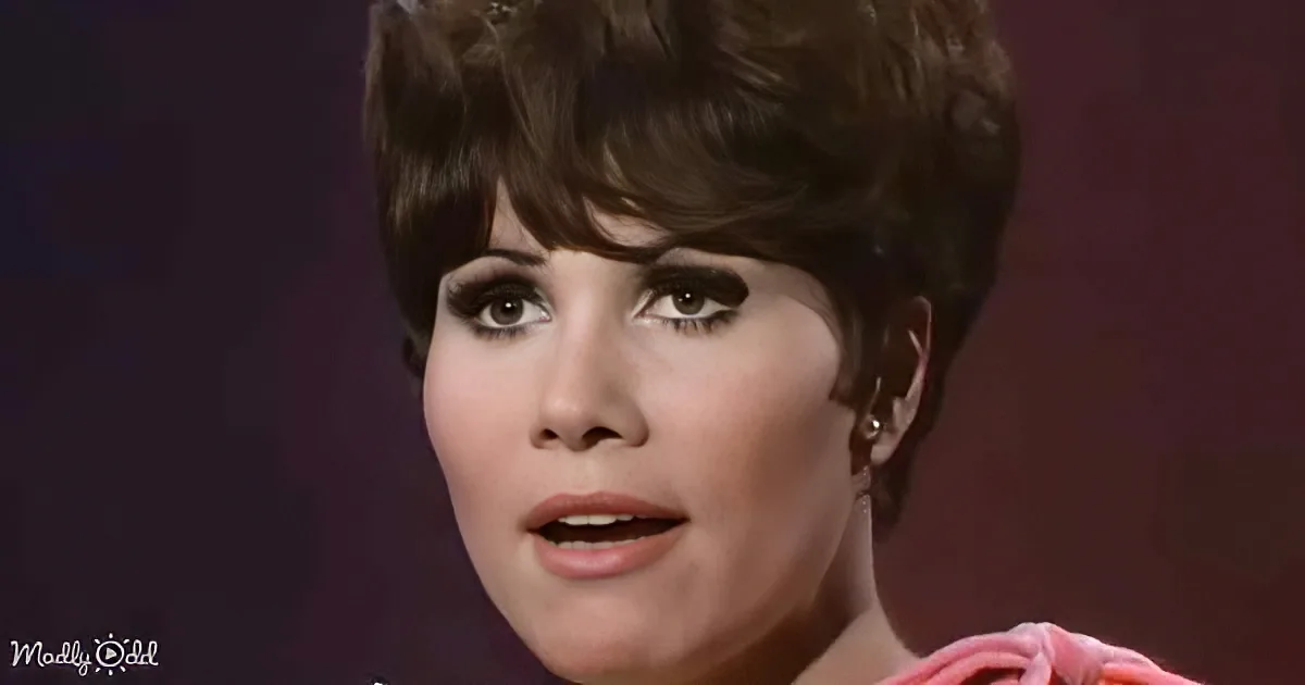A woman with short, styled hair and makeup, wearing a pink top, appears to be speaking or singing.