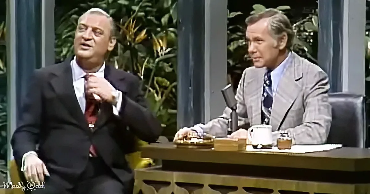A man in a suit adjusts his tie while another man in a suit sits at a desk with a microphone and coffee mug.