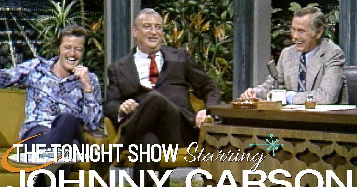 A man in a suit and red tie is laughing, sitting on a yellow couch, with another man in a patterned shirt and a third man...