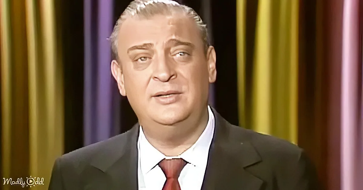 A man in a suit and tie stands in front of a colorful curtain backdrop.