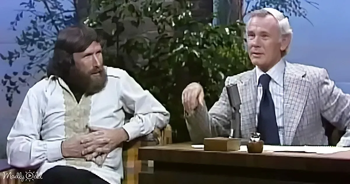A man with a beard sits in a chair, hands clasped, while another man in a suit gestures with his hand.