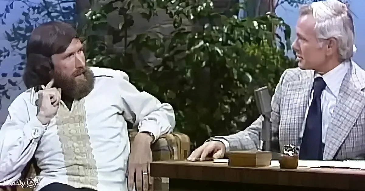 A man with a beard sits on a couch, engaged in conversation with a man in a suit at a desk.