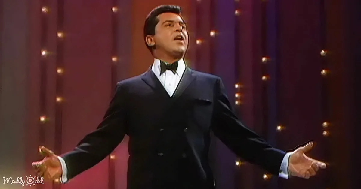 A man in a black suit and bow tie sings passionately on stage with arms outstretched.