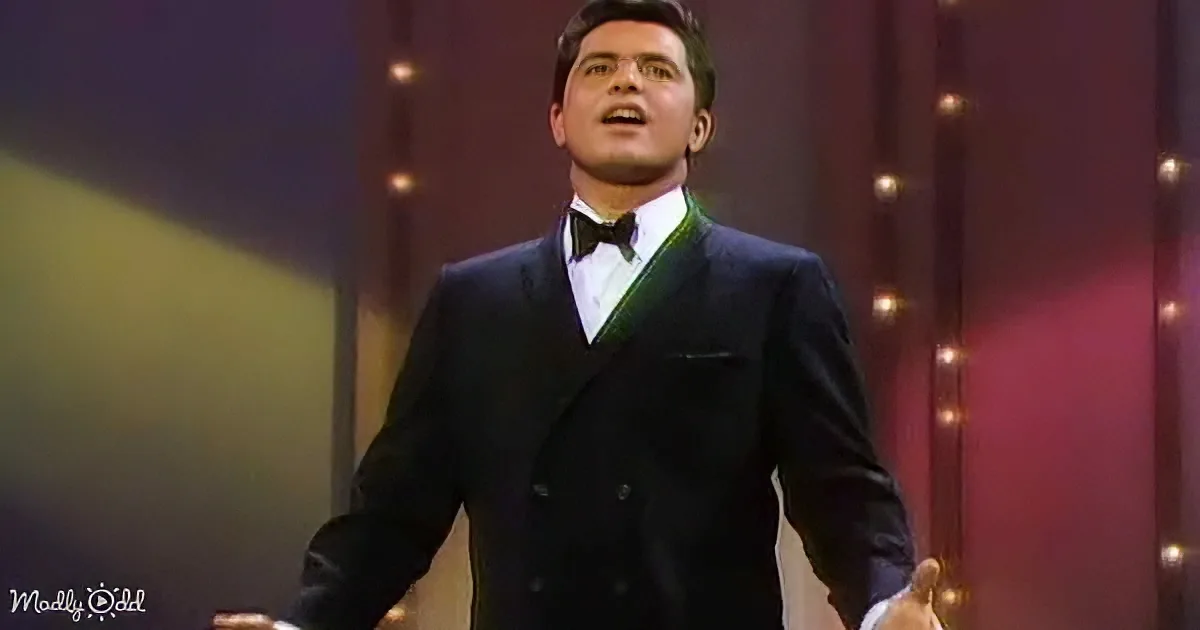 A man in a black suit and bow tie stands on stage, singing with a microphone in hand.