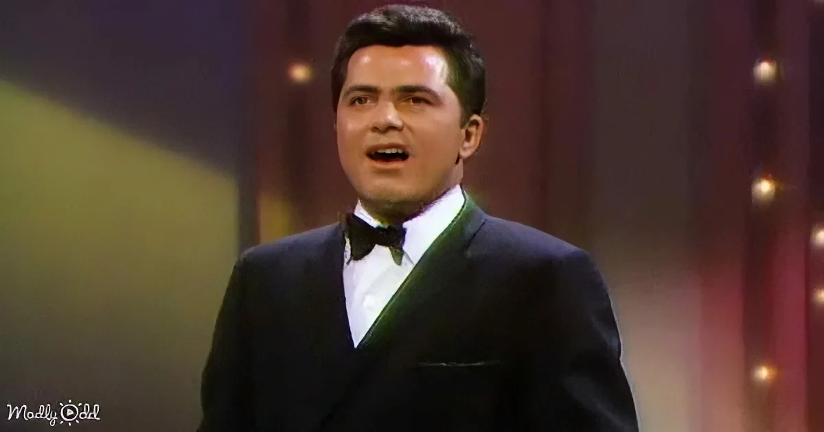 A man in a dark suit and bow tie sings on stage with a blurred background.
