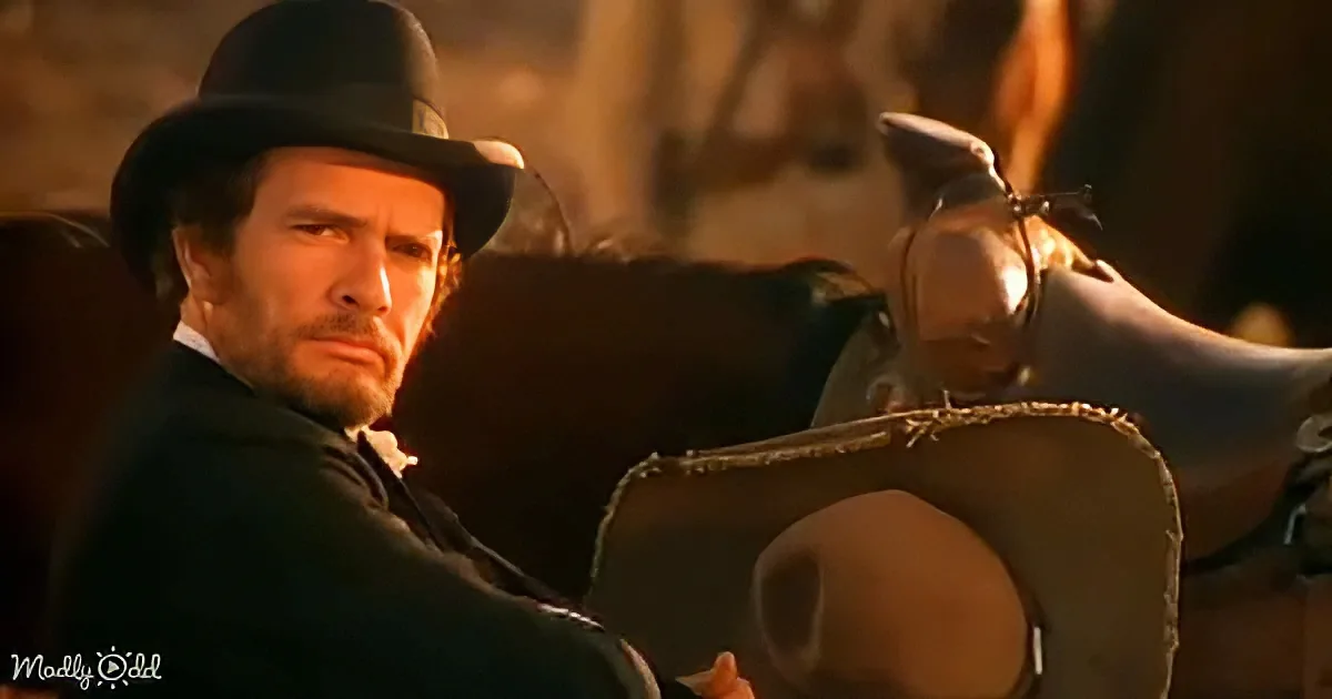 A man in a cowboy hat and suit leans against a horse, holding a gun, in a dimly lit scene.