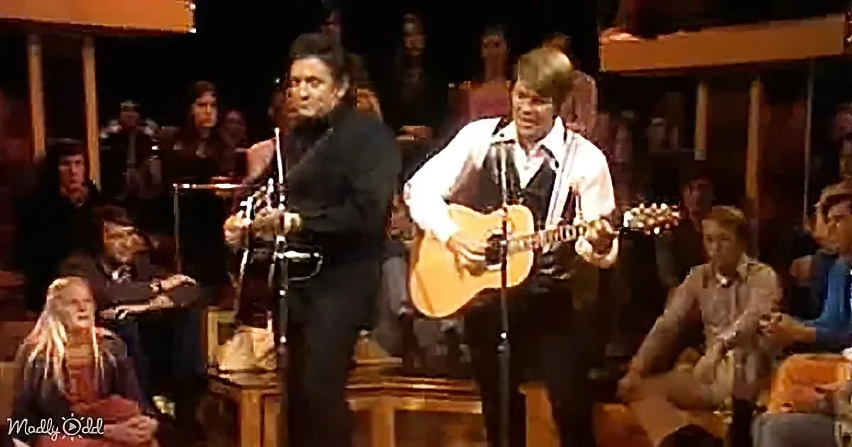 A man in black plays a fiddle while another man in white plays guitar on stage with an audience and orchestra behind them.