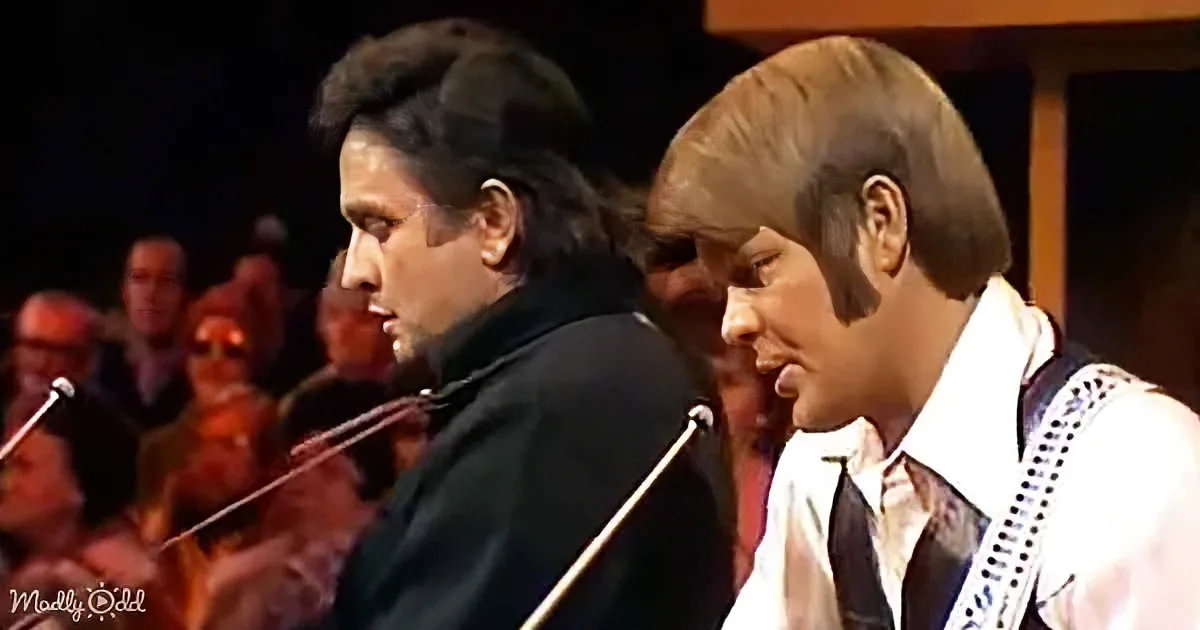Two men, one in black, the other in white, singing into microphones on stage with an audience in the background.