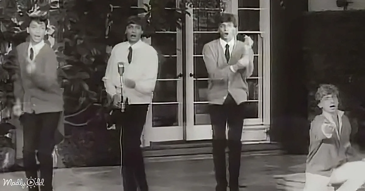Four men in suits sing on stage, one in a white shirt and tie, another in a suit jacket, and a third in a suit.