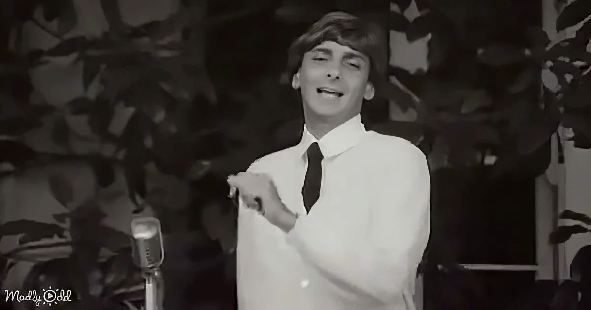 A man in a white shirt and tie smiles, pointing, with a microphone and plants in the background.