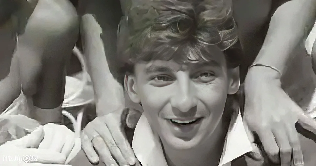 A smiling young man in a white shirt surrounded by others with hands on his shoulders.