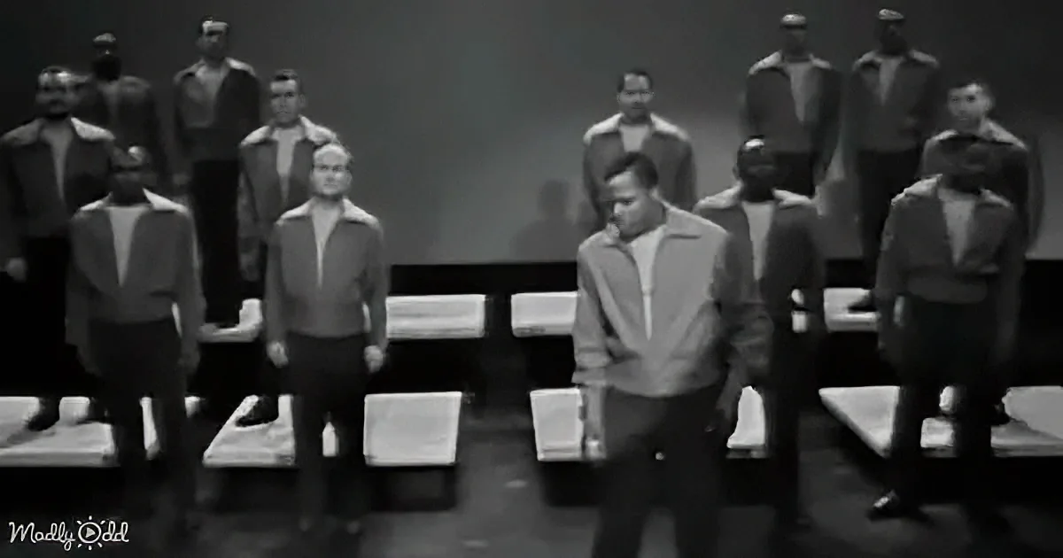 A man in a light jacket stands in front of a group of uniformed individuals in a dimly lit setting.