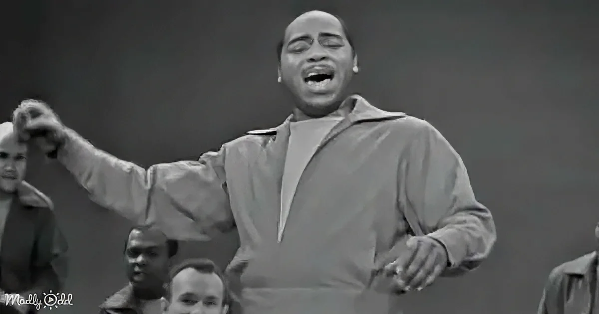 A man in a light-colored jacket raises his fist, singing passionately, with a crowd in the background.