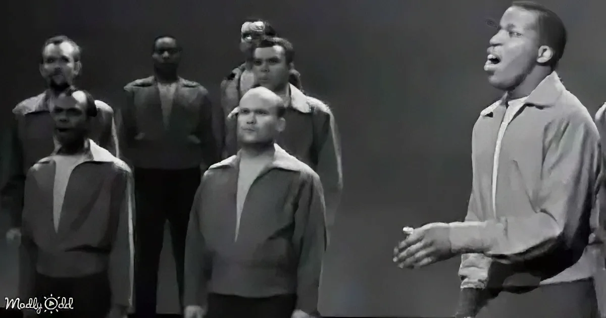 A man in a light jacket gestures while singing, surrounded by a group of men in darker attire.