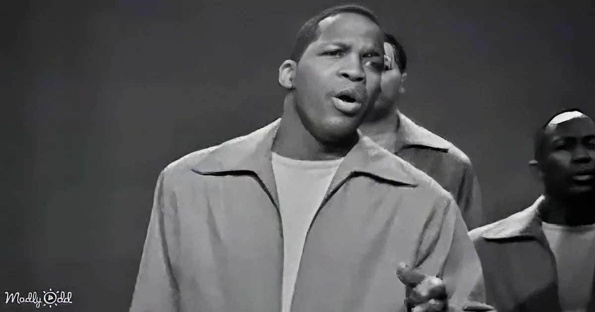 A man in a light-colored jacket sings passionately, with another person partially visible behind him.