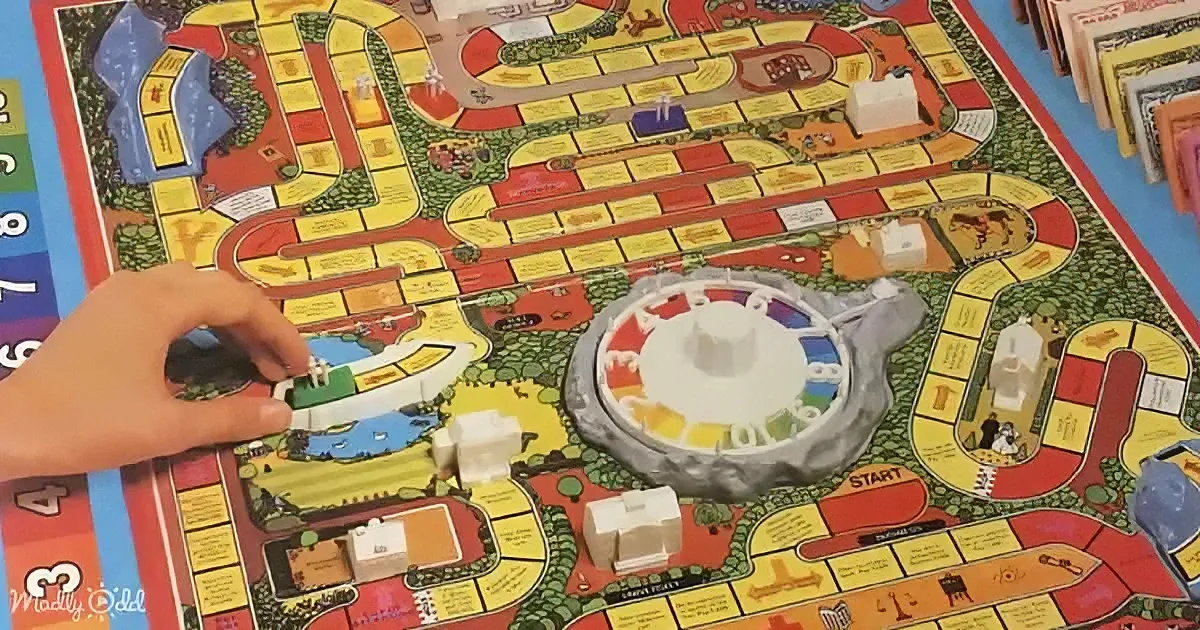 A hand moves a game piece on a colorful board game with various paths and spaces.
