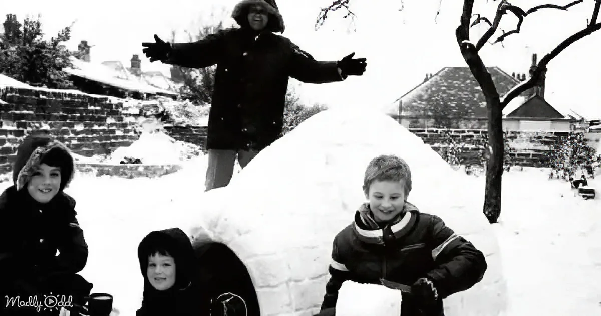 A person stands atop a snow igloo, arms outstretched, with three children in winter coats around it.