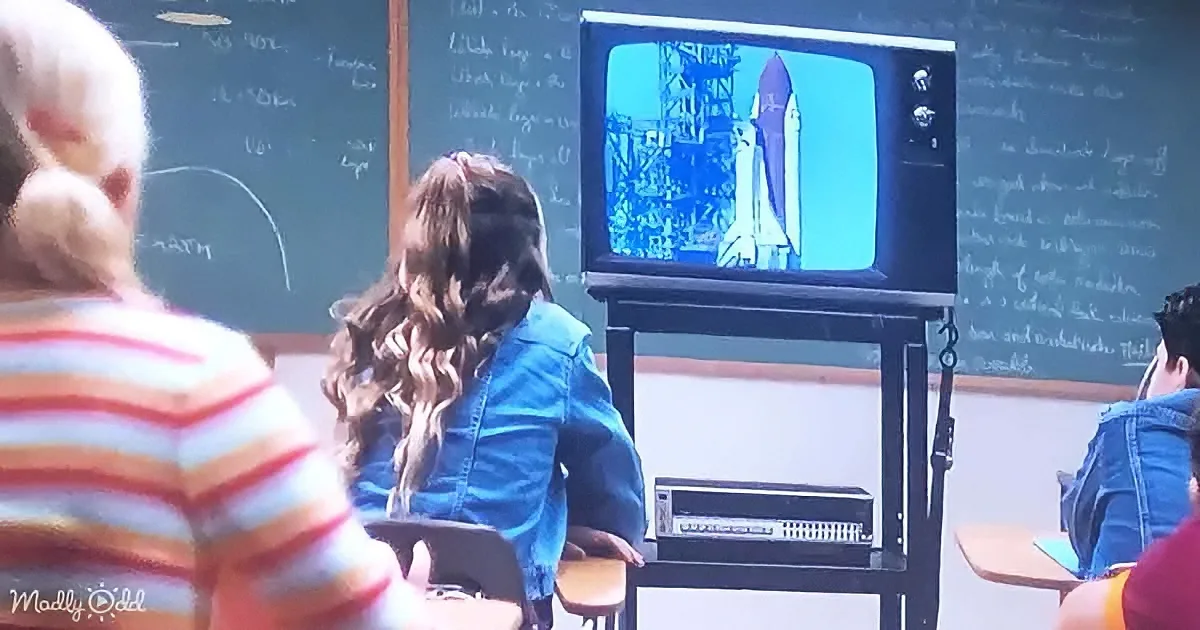 A young girl in a blue jacket stands in front of a classroom, facing a TV showing a space shuttle launch.