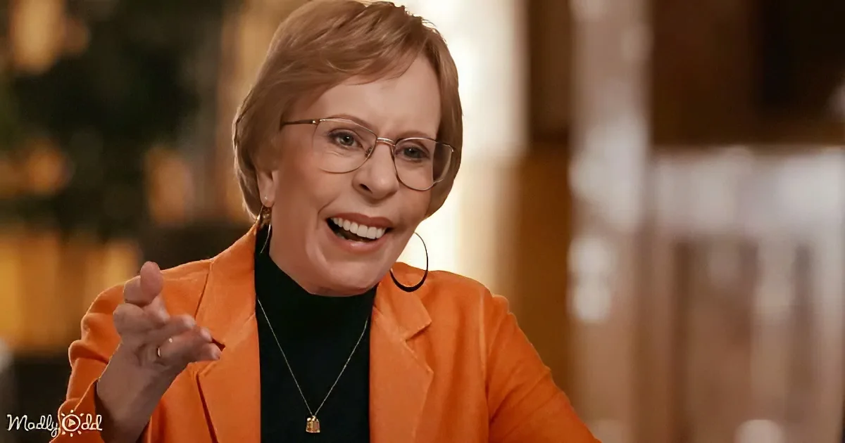 A smiling woman in an orange blazer and glasses gestures with her right hand.
