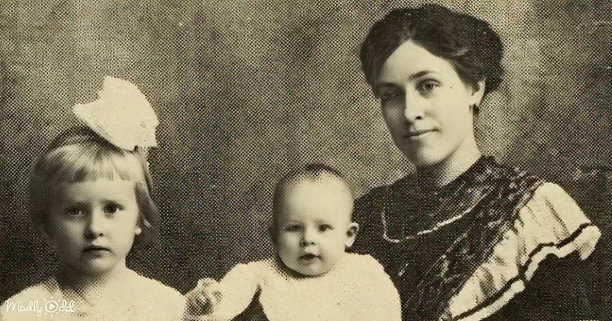 A woman holds a baby while another child stands beside them, all dressed formally, in a vintage portrait.